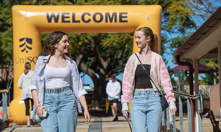 Two young women walk and talk under a yellow "WELCOME" archway outdoors, with trees and other people visible in the background.