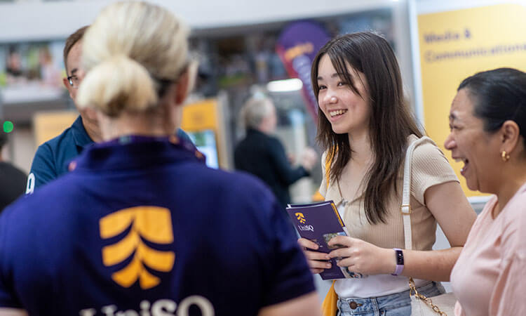 A young woman holding a notebook smiles and talks with two women at a UniSQ event, with informational booths visible in the background.
