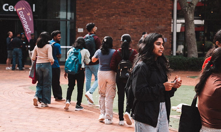 A group of students, some walking and some standing in conversation, are outside a building with brick walls and glass windows.