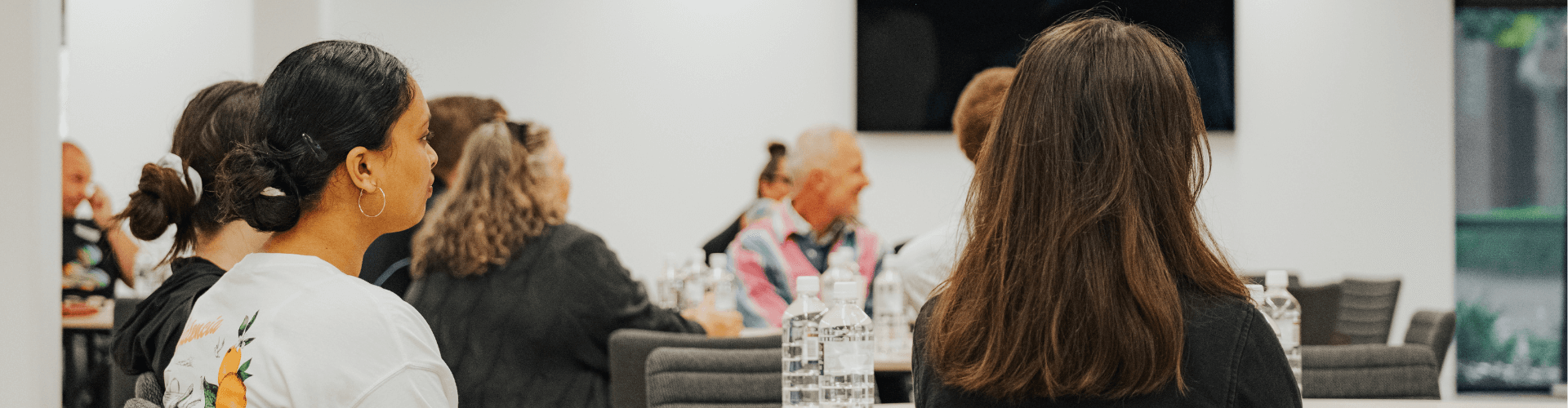 People sit at tables in a modern conference room, facing forward, with water bottles and notepads in front of them.