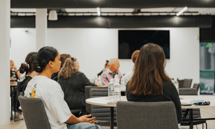 People seated at round tables in a modern conference room, facing forward and listening to a presentation.