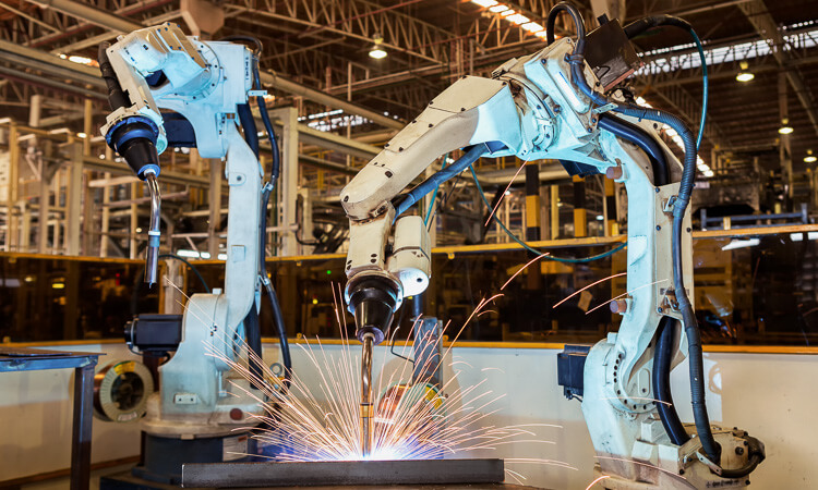 Two industrial robotic arms weld metal on an assembly line in a factory, producing bright sparks.