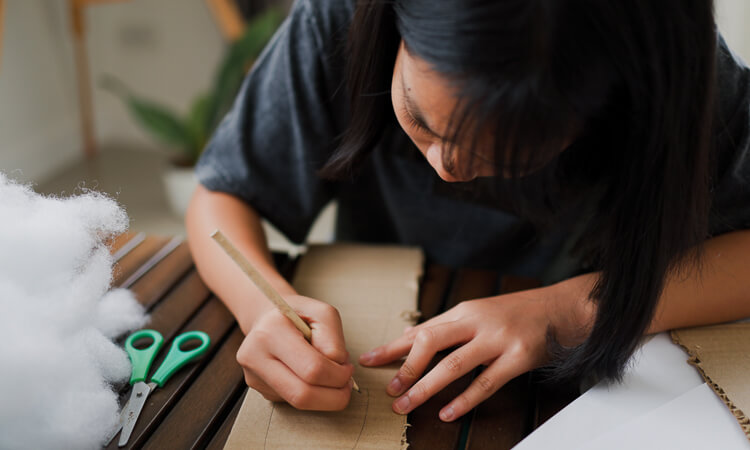 A person draws on a piece of cardboard with a pencil at a table. Scissors, polyester stuffing, and a plant are also visible nearby.