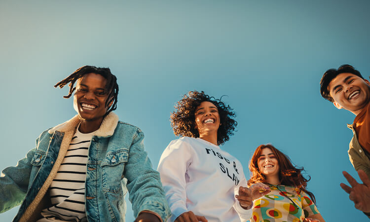 Four young adults stand outdoors, smiling and looking down at the camera against a clear blue sky.