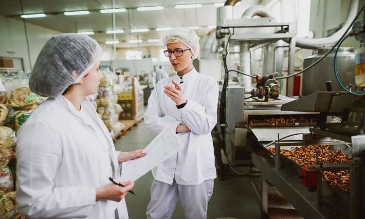 Two workers in white lab coats and hairnets discuss a clipboard in a food processing facility with machinery and packaged products in the background.