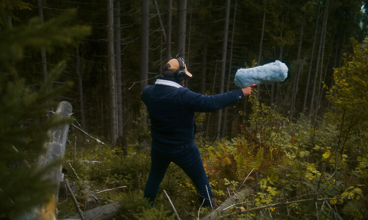 A person wearing headphones holds a boom microphone in a forest, surrounded by trees and vegetation.