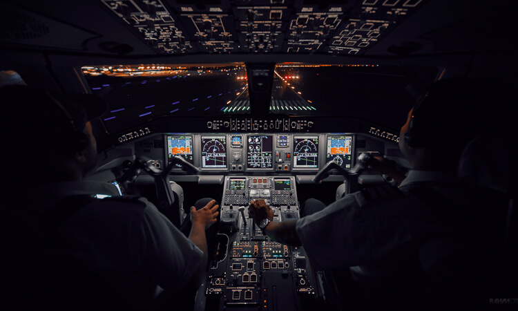 Two pilots in a dimly lit cockpit prepare to land an aircraft at night, with runway lights and illuminated instrument panels visible.