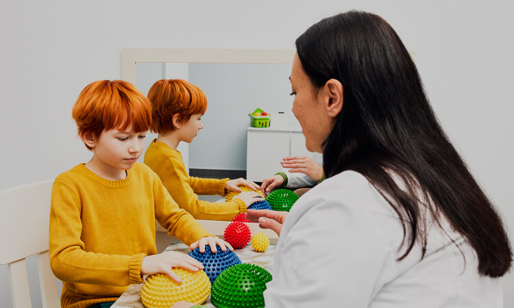 A child and an adult sit at a table with textured sensory toys in front of a mirror, engaging in a tactile activity.