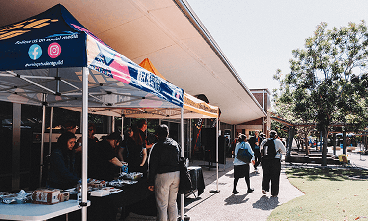 People gather under outdoor tents with tables set up for an event on a sunny day near a building and trees.