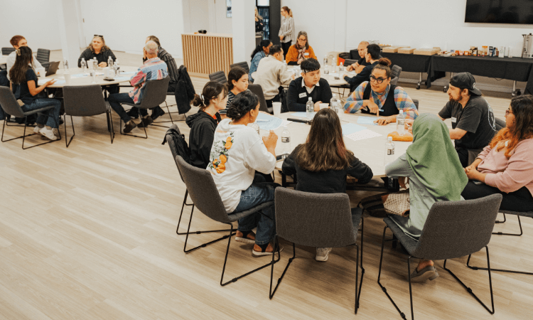 Groups of students sit around round tables in a modern meeting room, engaged in conversation and activities, with drinks and papers on the tables.