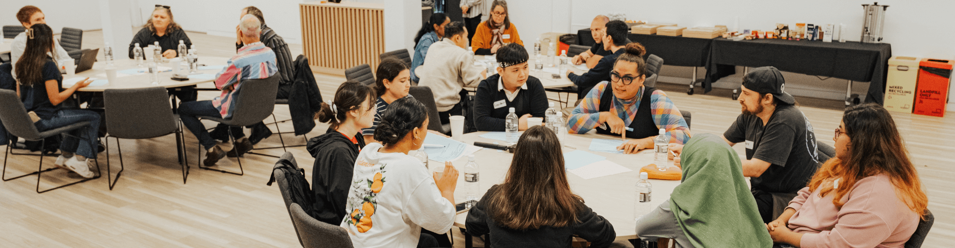Students sit in groups around round tables in a bright room, engaged in discussion. Drinks, papers, and name tags are visible on the tables. A refreshment table is in the background.