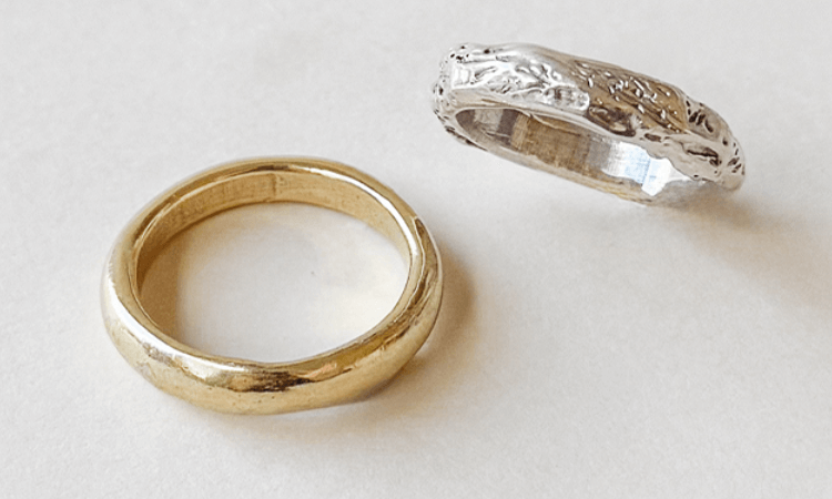 A gold ring with a smooth surface next to a silver ring with an intricate, textured design, both resting on a white background.