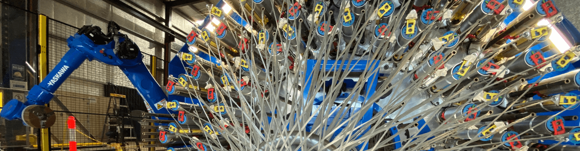 A robotic arm operates near a large circular machine with multiple cables and connectors inside an industrial facility.