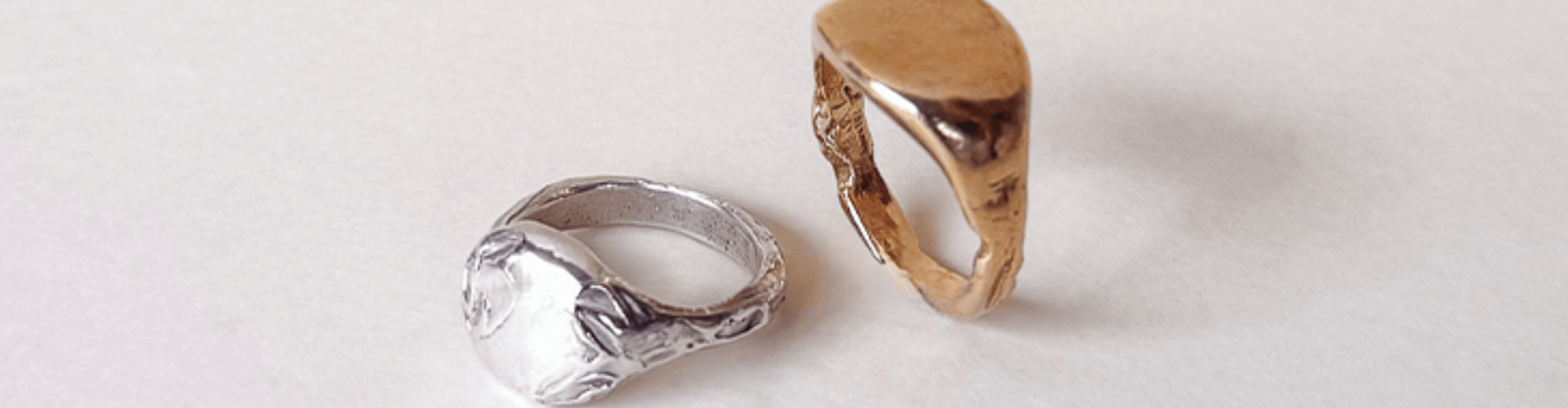A silver ring and a gold ring, both with irregular, organic shapes, are placed side by side on a plain, light-colored surface.