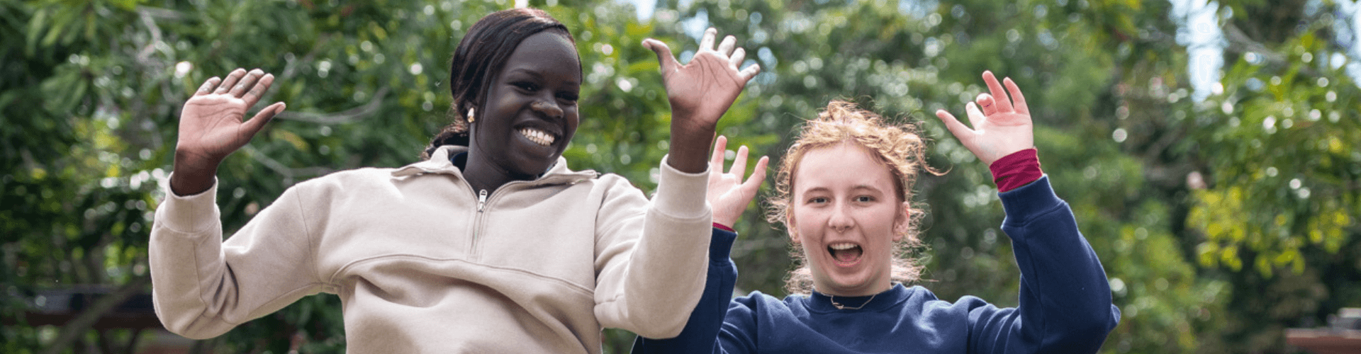 Two young women outdoors smile and raise their hands in the air, with green trees in the background.