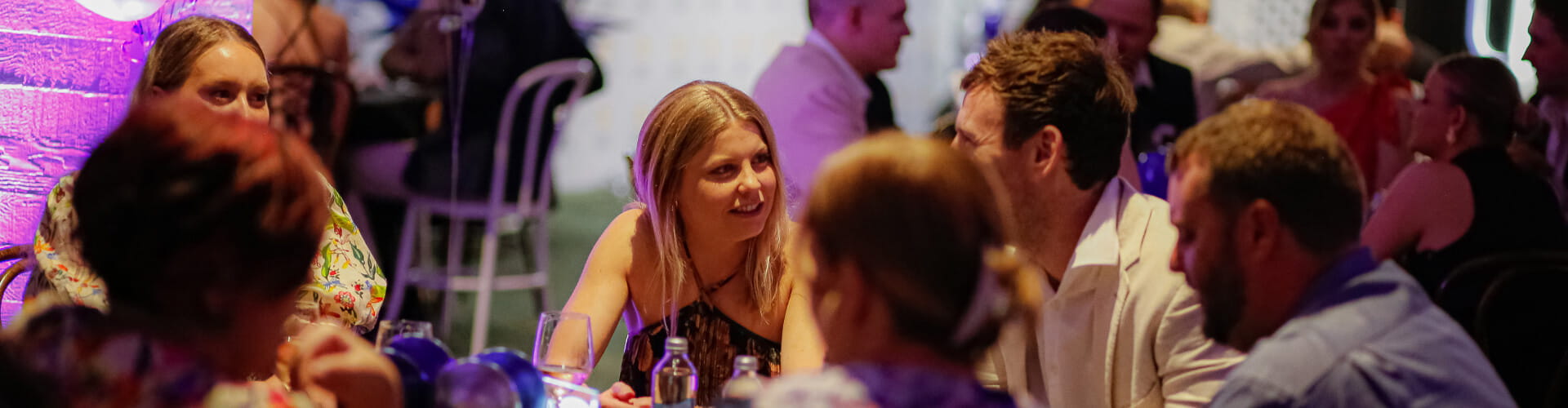 A group of people sit around a table at an indoor event, engaged in conversation with drinks in front of them.