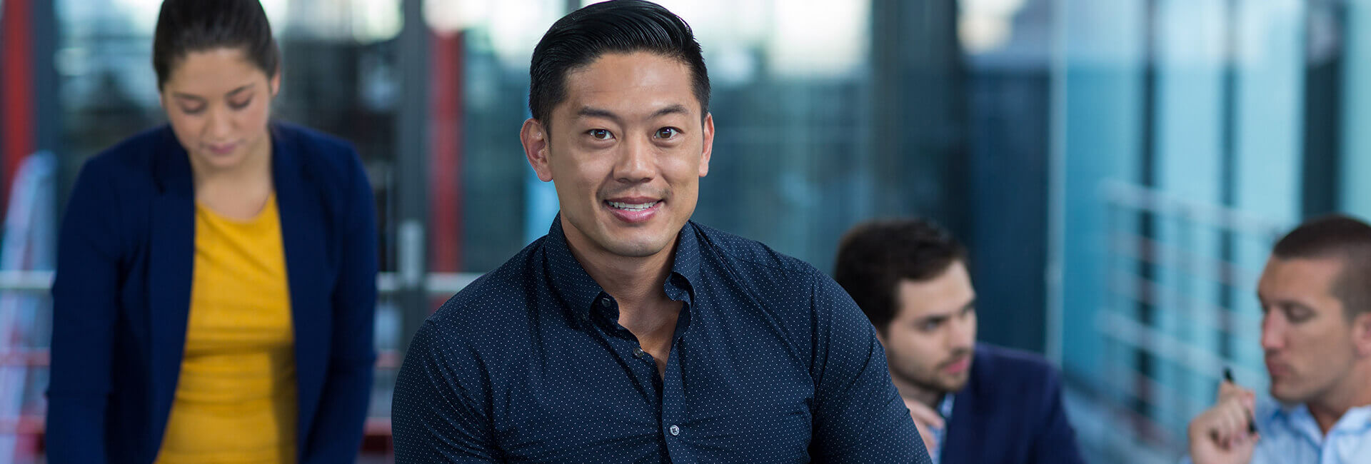 A man in a dark shirt sits and smiles at the camera, while three colleagues hold a discussion in a modern office setting in the background.