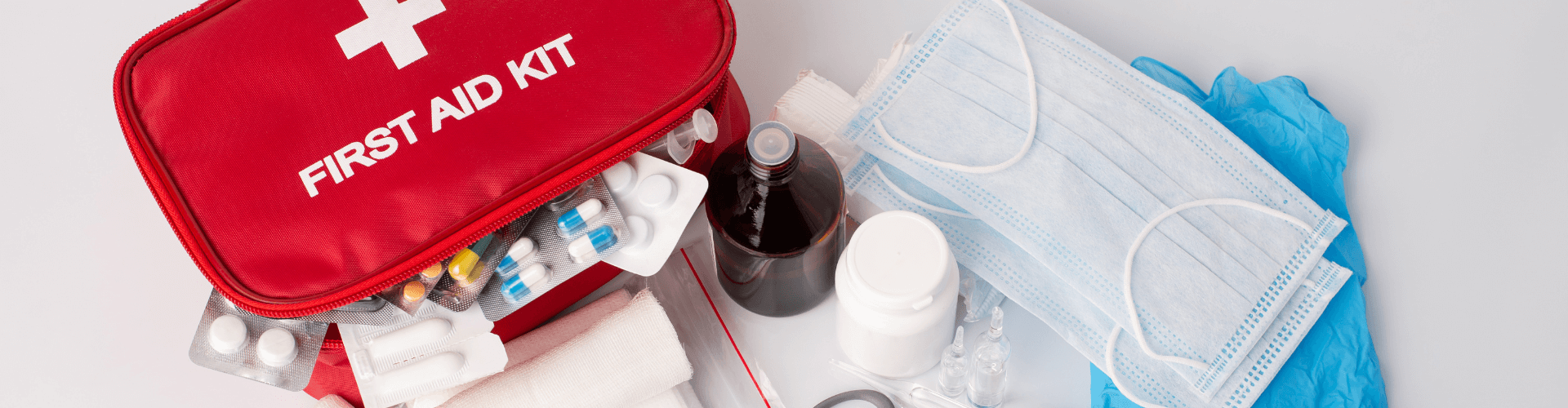 Open first aid kit with blister packs, medicine bottles, bandages, medical masks, and blue gloves displayed on a white surface.