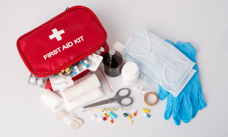 A red first aid kit with medical supplies including pills, bandages, scissors, gloves, face masks, adhesive tape, and medicine bottles arranged on a white surface.