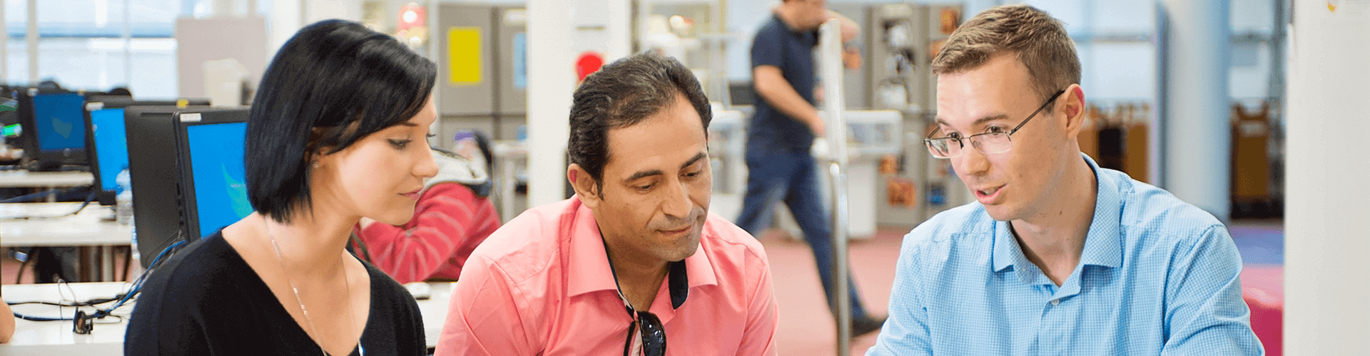 Three adults sit together at a desk in an office, looking at papers or a screen, while another person walks in the background.
