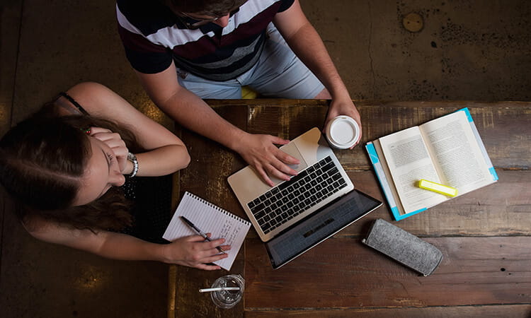 Two people sit at a wooden table with a laptop, open book, notepad, highlighter, cup of coffee, and glass of water, viewed from above.