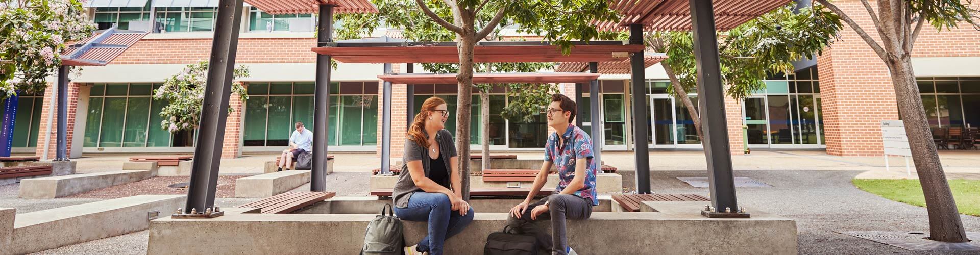 Two people sit and talk on benches under a pergola in an outdoor campus area with trees, brick buildings, and large windows in the background.