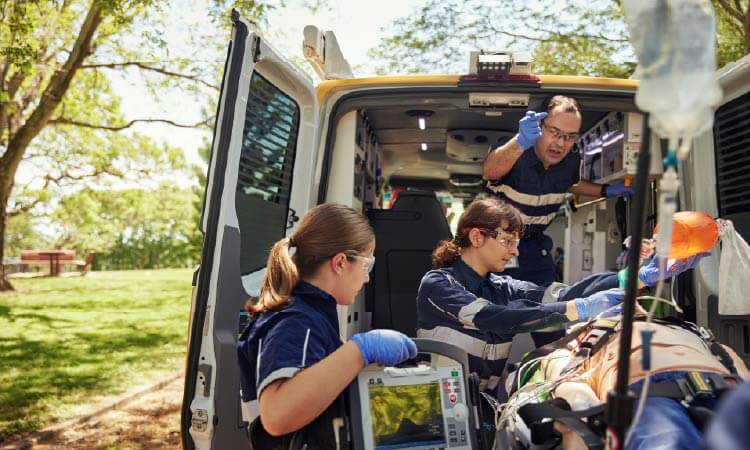 Paramedics attend to a patient on a stretcher outside an ambulance, using medical equipment, with one paramedic communicating on a radio.