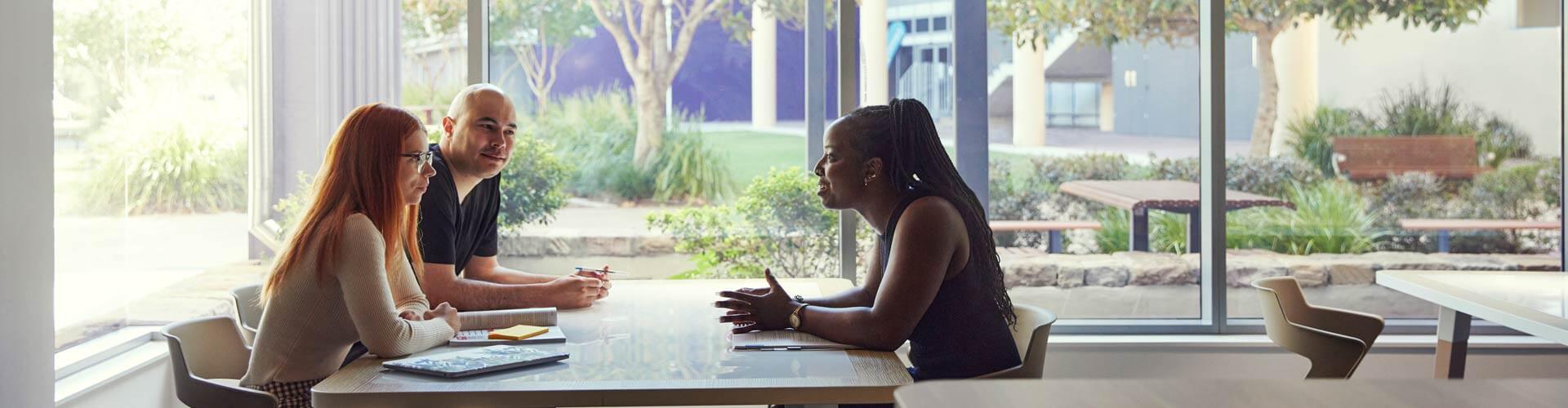 Three people sit at a table in a bright room with large windows, engaged in conversation with notebooks and laptops in front of them.