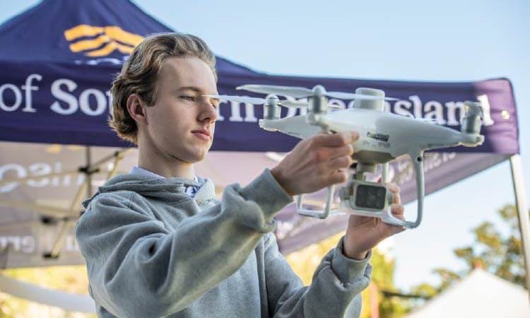 A young person in a gray hoodie holds and inspects a white drone outdoors, with a tent and trees visible in the background.