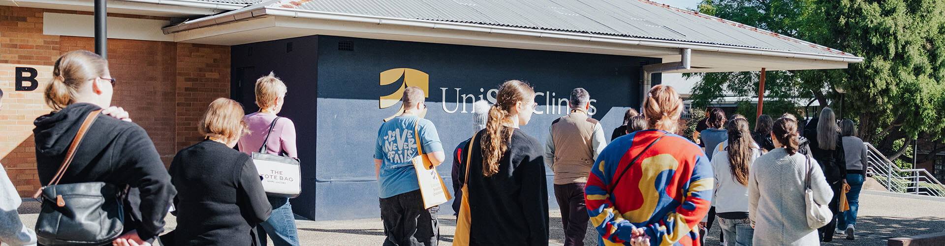 A group of people walk toward a building with a “UniSQ Clinics” sign on the wall, under a clear sky.