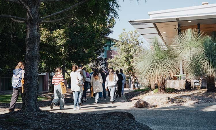 A group of people walk together along a sidewalk near trees and a modern building on a sunny day.