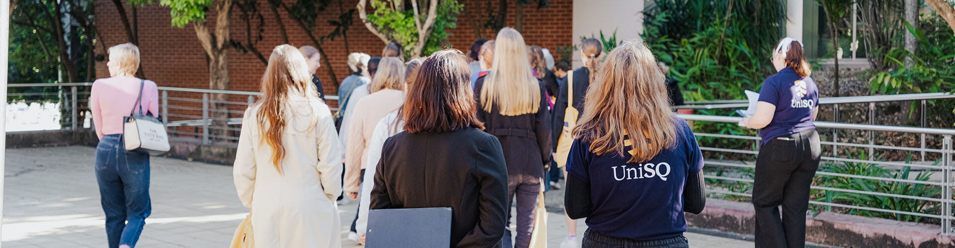 A group of people walks outdoors on a paved path near a building and greenery; one person wears a shirt with "UniSQ" on the back.
