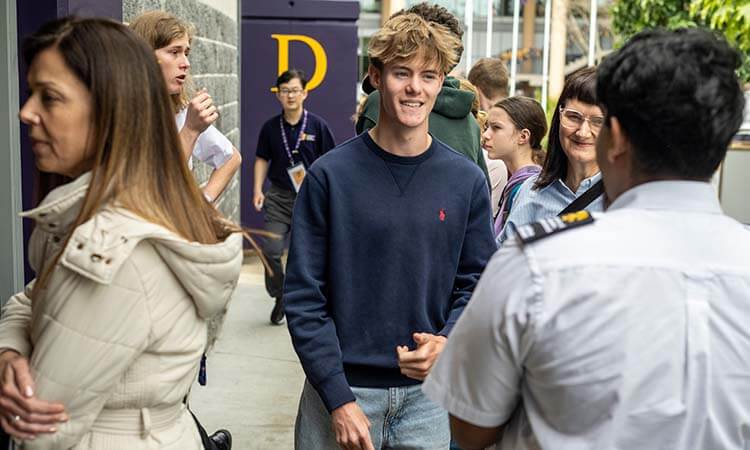 A group of people, including a young man in a navy sweatshirt and a person in a uniform, stand and talk outside near a building.