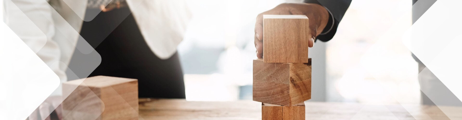 Two people stack wooden blocks on a table, with one hand adjusting the top block. The background is bright and out of focus.