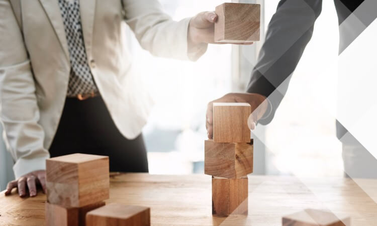 Two people in business attire stack wooden blocks on a table, suggesting teamwork or collaboration in a professional setting.