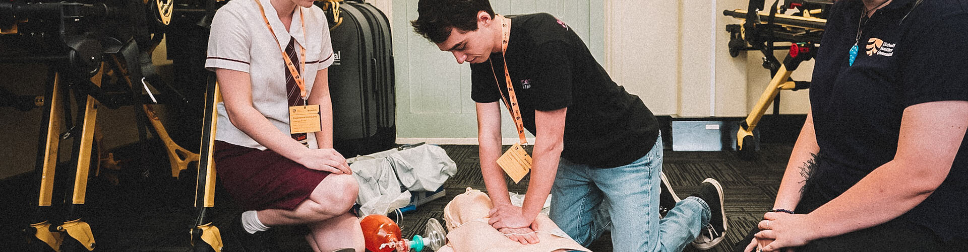 Three people practice CPR on a medical dummy; one performs chest compressions while two others observe. Medical equipment is visible in the background.