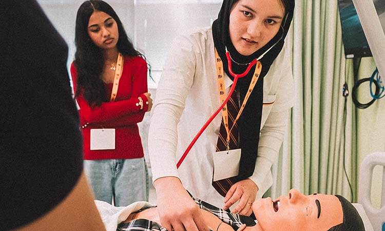 Two students practice a medical procedure on a mannequin, with one using a stethoscope while the other observes in the background.
