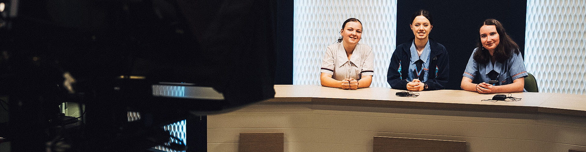 Three people in uniforms sit behind a news desk in a studio, facing the camera with microphones placed on the desk in front of them.