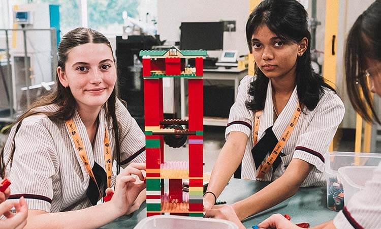 Two students in school uniforms build a tall structure with colorful plastic bricks at a classroom table, looking toward the camera.