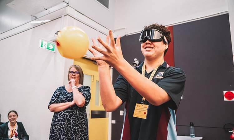 A person wearing goggles tries to catch a yellow balloon indoors while two people look on, one sitting and one standing with her hand on her chin.