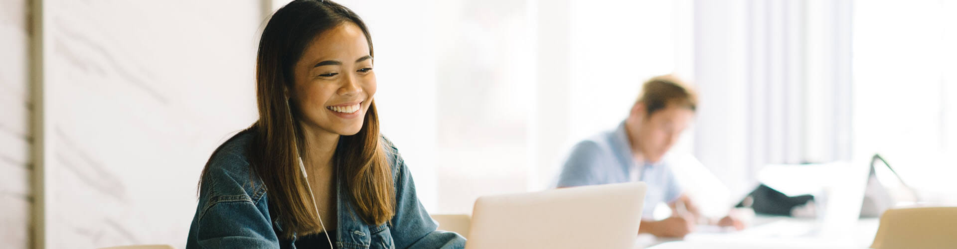 A young woman wearing headphones and a denim jacket smiles while using a laptop at a desk; another person works in the background.