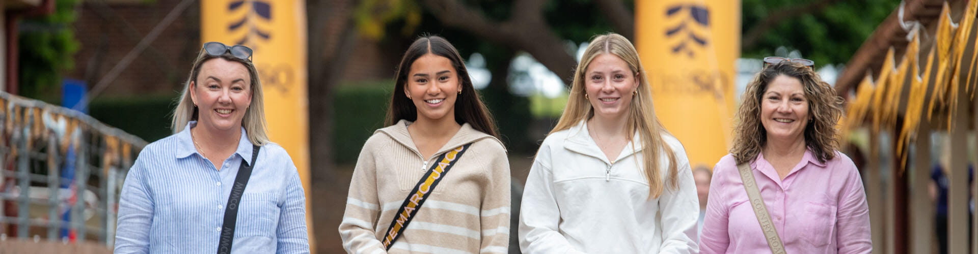 Four women stand side by side outdoors, smiling at the camera, with yellow banners and trees visible in the background.