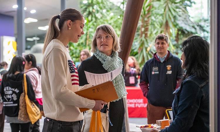 Several people are gathered indoors at an information booth, with one woman holding documents and speaking to another woman while others look on.