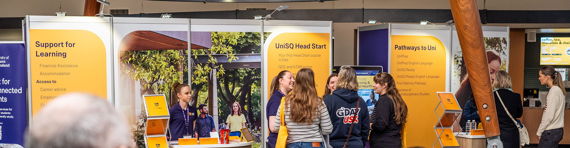 People interact at an information booth with banners about learning support and university pathways in a well-lit indoor setting.
