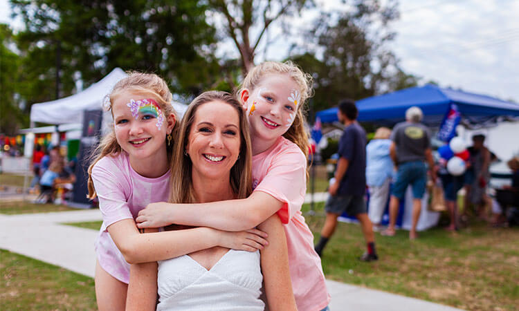 A woman smiles with two young girls who have painted faces, outdoors at a fair with tents and people in the background.