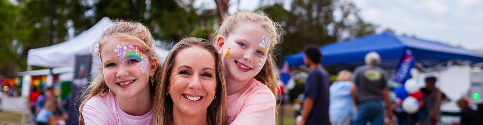 A woman smiles with two young girls who have face paint, posing outdoors at a community event with tents and people in the background.