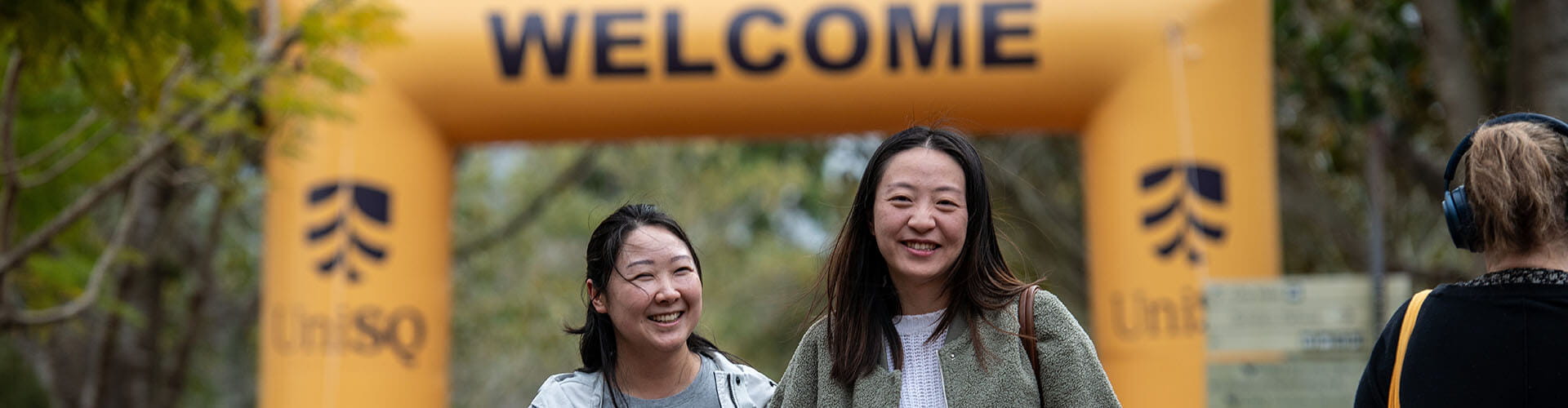 Two women smiling and walking outdoors in front of a large yellow "WELCOME" banner with trees in the background.