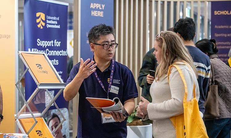 A university staff member speaks to a student at an information booth for the University of Southern Queensland, with brochures and banners displayed in the background.