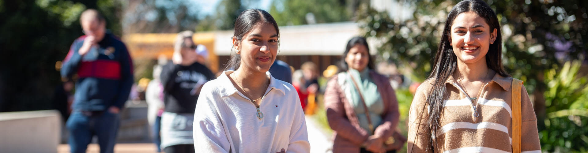 Two young women walk outdoors on a sunny day, with several people and greenery visible in the background.