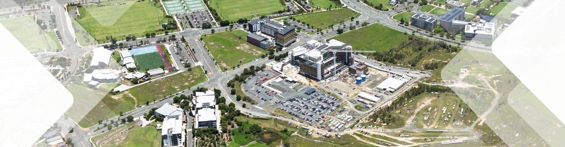 Aerial view of a large construction site surrounded by parking lots, roads, green fields, and multiple buildings in an urban area.